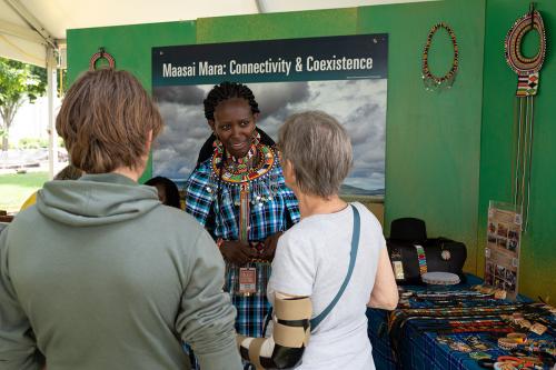 A woman in traditional Kenyan dress speaks to two individuals with their backs turned in an outdoor pop-up. Behind her is a sign that reads “Maasai Mara: Connectivity & Coexistence” and there are tables behind her littered with beaded designs. 
