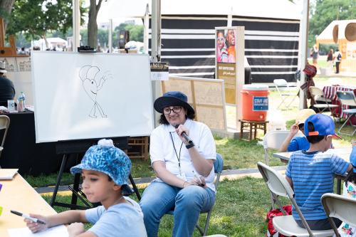 A woman, seated holding a microphone, smiles looking at the kids drawing at tables on either side of her. Behind her, a white board with a drawing of a smiling heart-shaped character with legs, an elephant trunk, and elephant ears. 