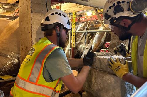 Two people in yellow safety vests, white hard hats, and work gloves, adjust stone bricks in a basement area. Yellow caution tape and silver ducts in the background. 