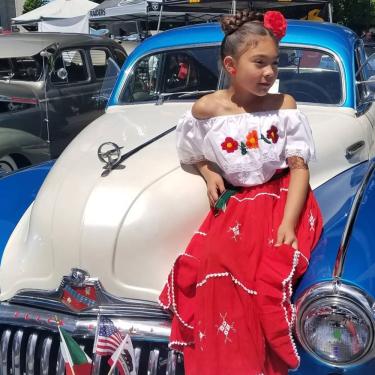 A young girl wearing Mexican floral blouse and long red skirt, with a red flower in her hair, poses sitting on the hood of a blue and white classic car. Three small flags stick up from the front bumper: Mexico, United States, and California.
