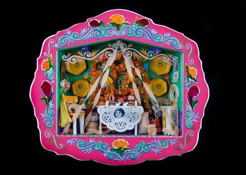 A small handheld shadow box with a bright pink frame with floral motifs in white and blue. Inside of the box is a diorama of a traditional Día de los Muertos/Day of the Dead domestic altar scene, including a table holding offerings of food, dishes, and pl