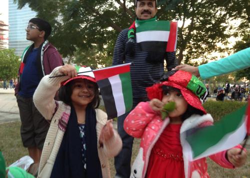 Young girls and adults hold up red, green, white, and black UAE flags, waving them in celebration. In the background are trees and two men. 