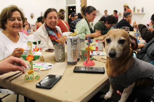 Two women, among a group sitting around a table painting sculptures, smile at a small dog, held up at table level and looking sternly at the camera.