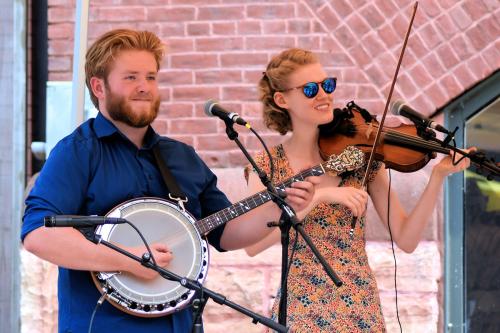 Two people perform onstage outside on a sunny day: a young man with blond hair and beard and blue dress shirt on banjo, and a young woman with blond hair, sunglasses, and a floral dress on fiddle.