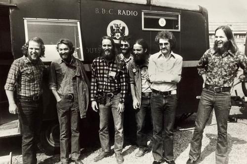 Seven men stand, most laughing, outside a trailer with BBC Radio painted on the side. They wear worn jeans and button-up shirts.