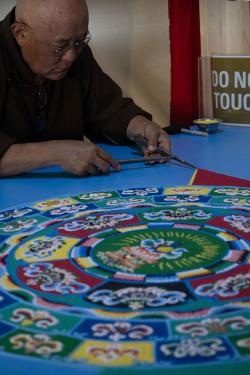 A man uses two tools to pour a yellow line of sand in concentric circles of blue, yellow, green, red, and pink flower designs.
