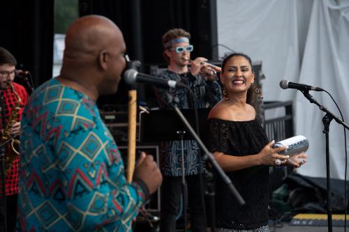 Four people on stage performing. In focus is a woman smiling, shaking a silver cylindrical percussion instrument.