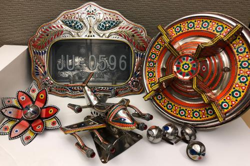 Painted chrom objects on a desk: a license plate with silver frame with a feathery, floral design. A hubcap with red, yellow, and black floral design. Silver bells. A hood ornament in the shape of an airplane, silver with red and yellow trim. A red and si