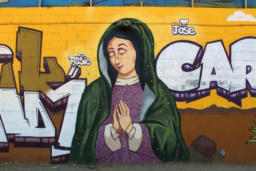 Mural on a brick wall featuring the virgin in purple dress and green shawl, with graffiti letters in purple, orange, and brown behind her.