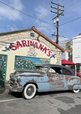 A pale blue classic car with rust spots parked on the street in front of a building.