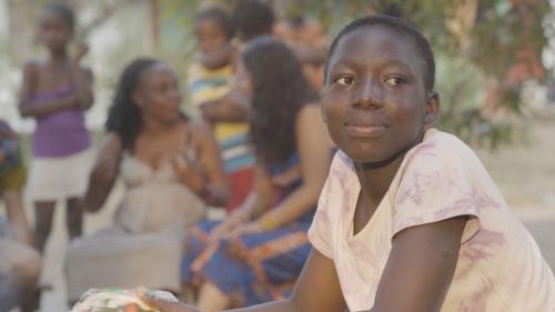Film still of a person sitting outside, looking into the distance, while others gather behind them chatting.