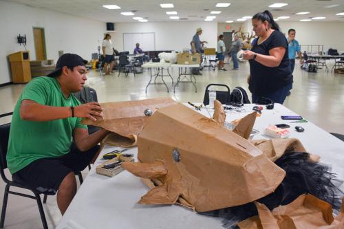 Seated at a table, a person constructs a costume piece from brown material. In the foreground, a buffalo head costume piece.