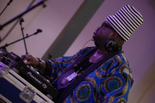 A man in a striped conical hat and blue floral jacket DJs on a turntable.
