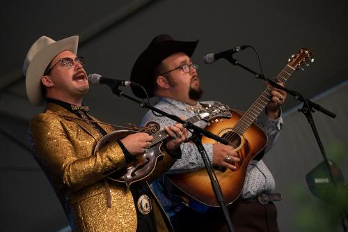 Two men perform on stage, one in gold jacket singing and playing mandolin. The other plays acoustic guitar.