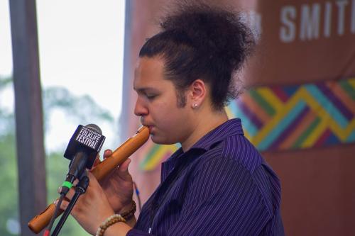 A young man with dark curly hair up in a bun closes his eyes as he plays a wooden flute on stage.