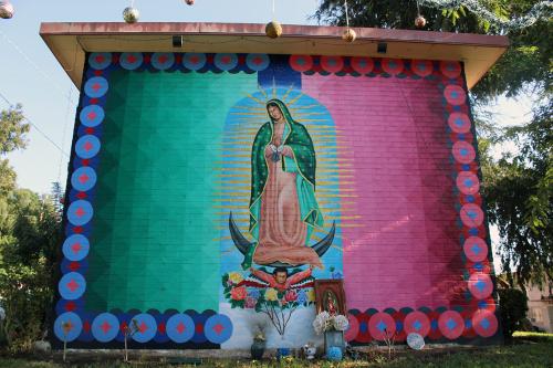 Mural on the side of a brick building, surrounded by trees. The painting features the virgin in shades of pink and teal.