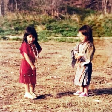 Two small kids with long dark hair and wearing dresses face each other, standing in dirt.