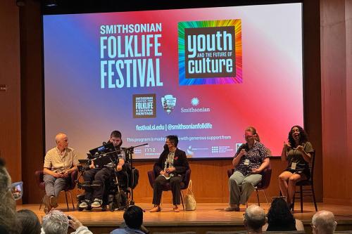 Five people sit on stage in front of an audience. On one side, three people are in conversation. On the other, one person is audio describing and the other is interpreting the conversation into sign language.