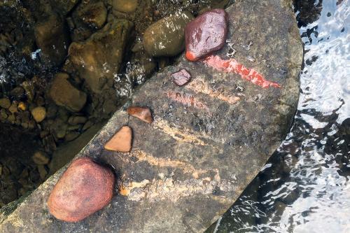 Five small stones, in a range of colors from red to orange, have each produced a streak of colors on a wet rock along a river bank.