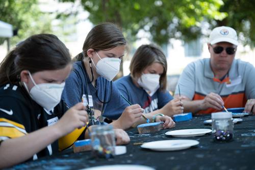 Four people sit at a table outside, painting palm-sized pieces of wood blue.