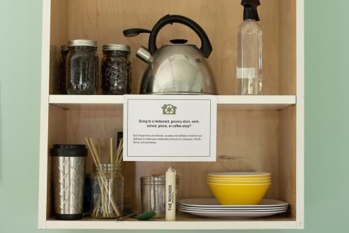 Close-up of a mock kitchen cabinat set against sea green wall, containing coffee beans in a jar, silver kettle, yellow and white dishes, bamboo straws, and other kitchenware. A sign reads: Going to a restaurant, grocery store, work, school, picnic, or cof