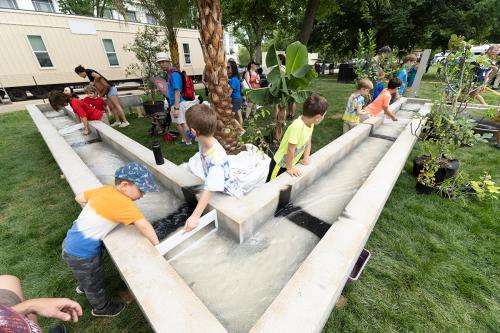 Kids surround the edges of an outdoor, elevated cement waterway with water flowing through it. In the background, palm and other plants surround the structure.