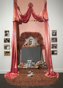 A large altar installation piece framed by two pink curtains and multiple framed black-and-white photographs on either side side, holding within a large mirror shrouded with a dark pink fluff. The altar holds multiple smaller shiny items, candles, photogr