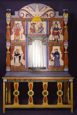 A large handmade altar with a protruding table-like base and a painted backboard consisting of five scenes depicting Catholic saints and angels, and a large rounded dove on the top. In the middle is a space  reserved for a statue to be placed at the cente