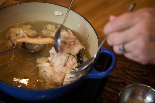 Closeup on a pot of chicken soup as it simmers on a stovetop. Using two spoons, someone is shredding the chicken.