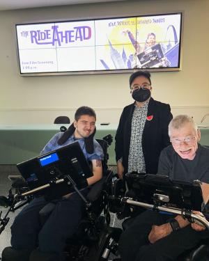 Three people pose, smiling, under an advertisement for The Ride Ahead on a screen above them. Two of them, a young man and an elder man, are in wheelchairs with communication devices attached. The third person stands between them, wearing a face mask.