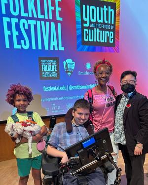 On stage under a projection advertising the Folklife Festival, four people smile and pose. A young child holds a small white dog in their arms.