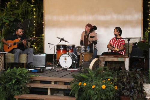 Three people play music on an outdoor stage: acoustic guitar, drum kit, and electric guitar.