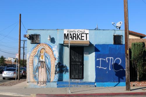 A blue corner commercial building with a sign above the door, Santa Cruz Market, and a painting of the virgin in blue and tan. Graffiti on the other side of the door reads ITC.
