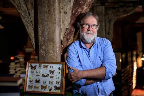 A white man with gray hair and beard, wearing glasses and blue dress shirt, poses next to a framed display of butterflies.