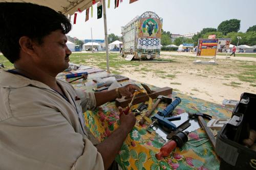 A man with dark skin sits at a table under a tent outdoors, cutting something with scissors. He has various tools in front of him. In the distance, the back of the painted truck parked on the grass.