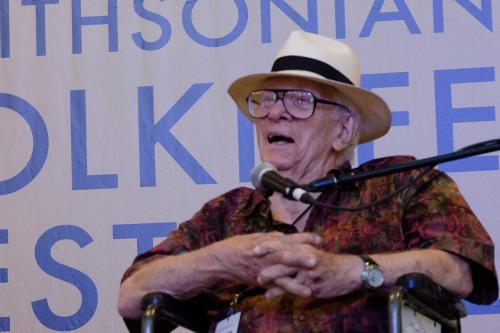 A much older Jim Morris sits on a Smithsonian Folklife Festival stage, speaking into a microphone. He wears glasses, a tan fedora, and a short-sleeved dress shirt.