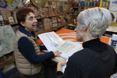 Two women talk over laminated pages of artwork. Art supplies stacked in the background.