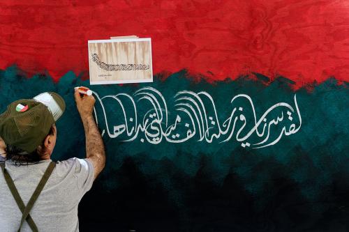 A man paints Arabic calligraphy on a red, green, and black painted-wooden wall, using a picture with the same calligraphy taped to the wall for guidance.