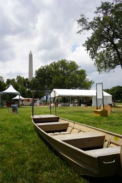 A simple, rectangular silver metal rowboat on the grass, with the Washington Monument, trees, and white tents in the background.