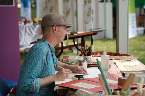 A man in a newsboy cap and floral turquoise shirt sits at a table outside, holding a calligraphy pen, surrounded by paper, books, and writing tools.