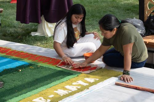 Julia Sánchez and Vicky Cabrera Sánchez at work on the alfombra, using a stencil to press red dyed sawdust onto the outline of the Maya woman weaving scene.
