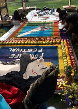 Behind a painted three-dimensional figure of Jesus Chris, a platform in the grass is designed with dyed sawdust and whole fruits, including apples, oranges, and pineapples. Several people sit and kneel to add to the design.