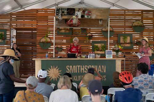 On a kitchen demonstration stage, a woman in red blouse and apron listens as someone in the audience asks a question. Above the chef, a mirror shows her kitchen counter and a pot of food on the stove.
