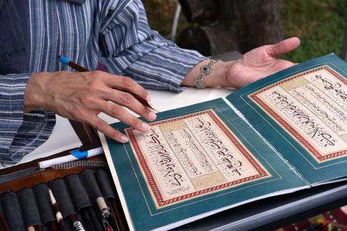 A person holding a reed stylus gestures toward an open book of Arabic calligraphy instruction.