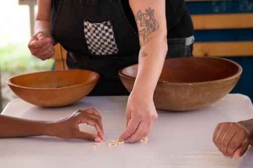 Under a white tent, a visitor uses a giant mortar and pestle with Elena Terry oversees. Other visitors sit at nearby tables, set with ears of corn and bowls of corn kernels.