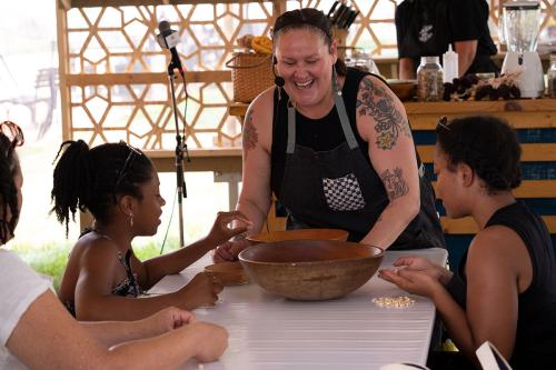 Around a table outdoors, a woman with bare, tattooed arms laughs as she handles wooden bowls. Others seated around the table hold kernels of corn.