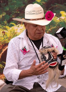 A man in white button-down shirt and white cowboy hat, both adorned with pink flowers, holds up a carved wooden face mask with red mouth and beige hair on the brow and chin.