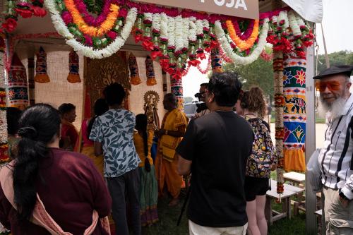 People crowd into a tent festooned with flower garlands and textiles with floral and elephant designs. Everyone is focused on someone at the back of tent, except for a man in orange-lensed glasses who peers directly at the camera.