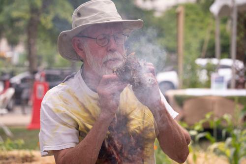 A man in a beige safari hat holds a dry, red-brown plant amidst a cloud of smoke.