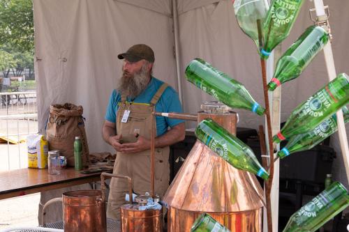 A man with big gray beard and tan overalls speaks from behind the copper pots and pipes of a still. In the foreground, a bottle tree: green glass bottles suspended upward from metal branches.